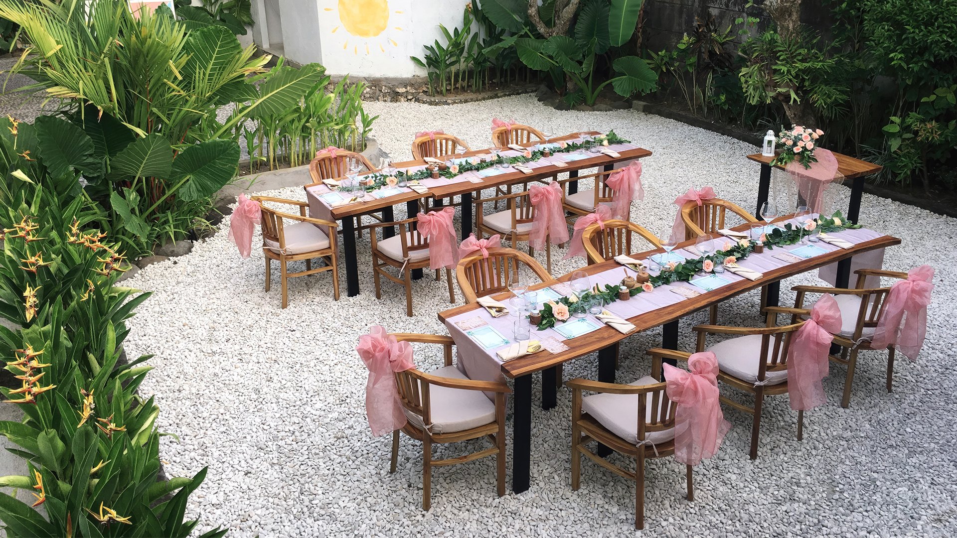Outdoor celebration tables set with flowers and pink sashes in a tropical garden